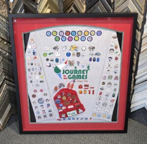 Custom shadow box frame displaying a Journey to the Games Italy '26 shirt covered with Olympic and winter sports pins, along with a commemorative beanie and memorabilia.