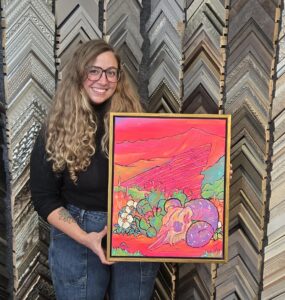 Smiling woman holding a vibrant Southwestern desert painting featuring bright pink and orange mountains, a ram skull, purple horns, flowering cacti, and colorful desert plants. The artwork is displayed in a sleek gold custom picture frame inside Frame de Art, surrounded by a wall of decorative frame samples.